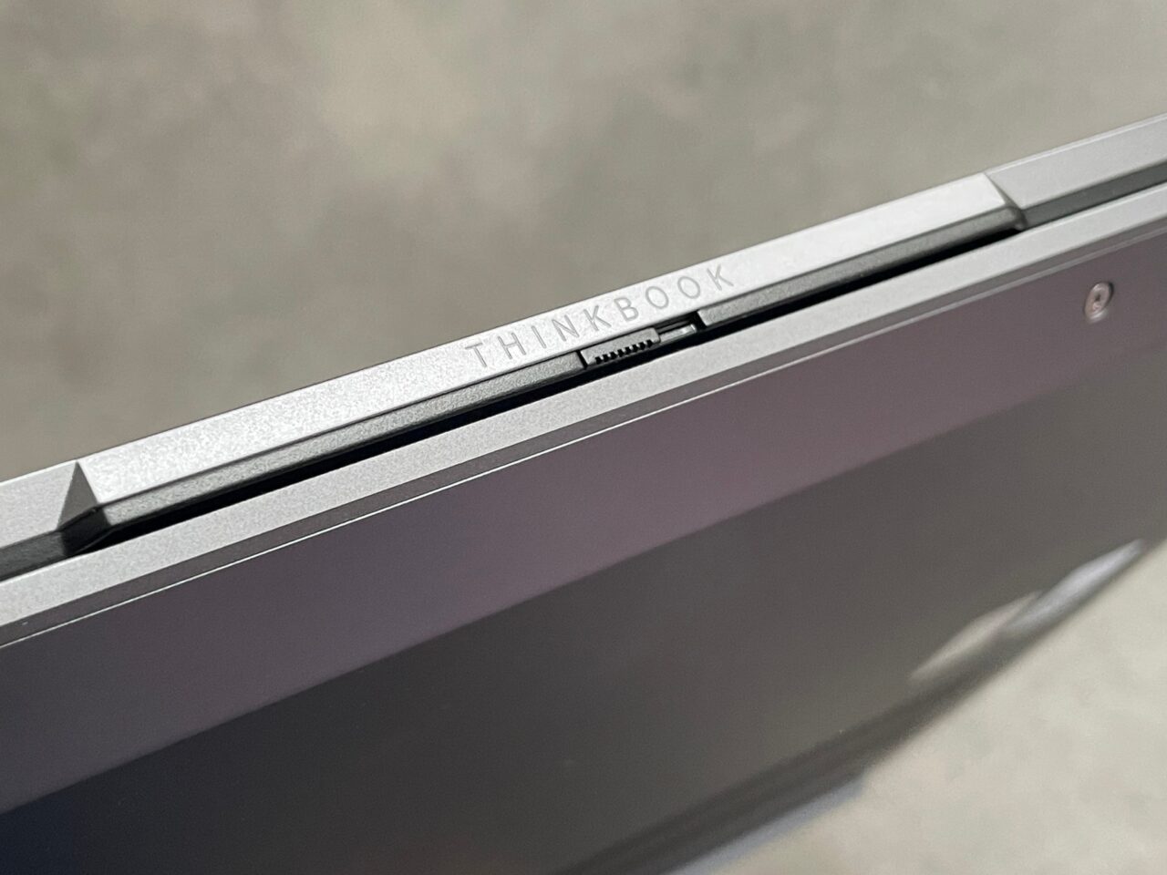 Zbliżenie na narożnik srebrnego laptopa z wygrawerowanym napisem "THINKBOOK" na zawiasie obok kamerki internetowej.