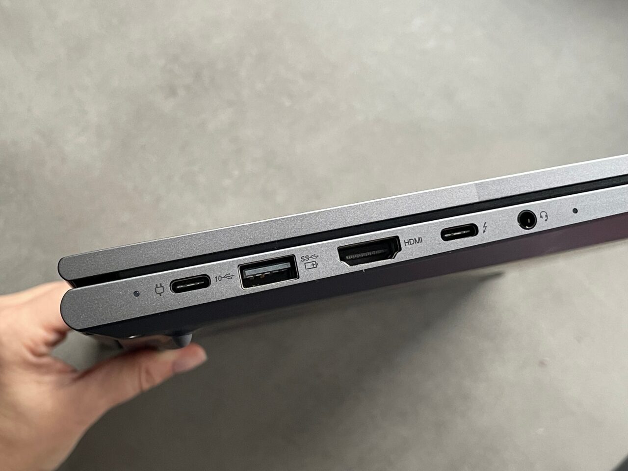 Bok laptopa trzymanego w dłoni z widocznymi portami: USB-C, USB-A, HDMI, gniazdo słuchawkowe i karta SD.