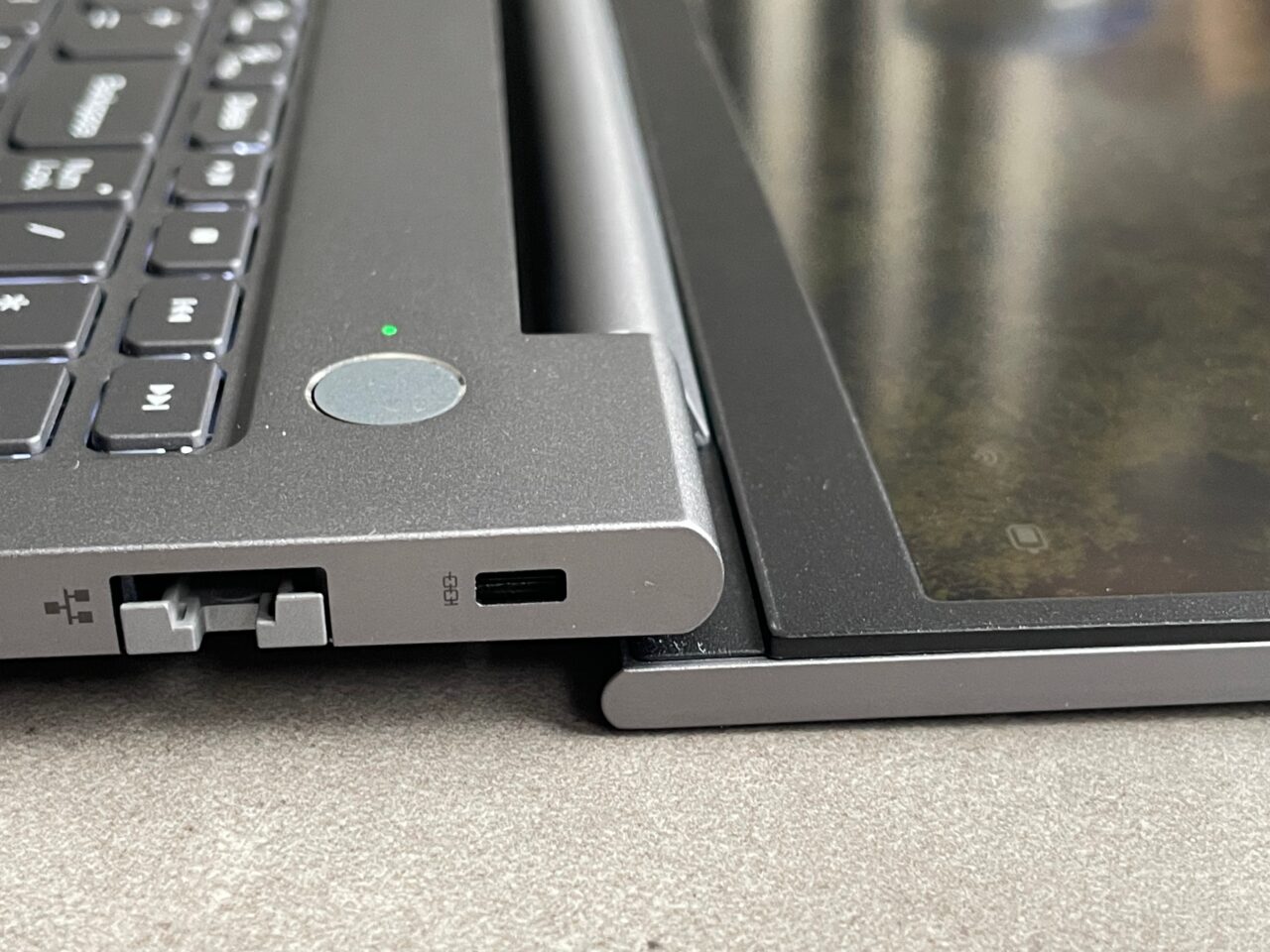Część laptopa z widocznym portem USB-C, przyciskiem zasilania i częścią klawiatury na pierwszym planie, oraz częściowo zamazanym ekranem laptopa.
