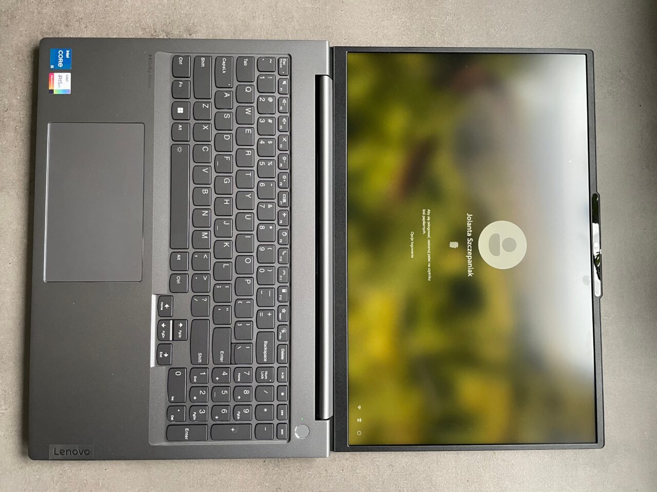 Otwarty laptop marki Lenovo na szarym blacie, z widoczną klawiaturą i ekranem logowania systemu Windows, rozmytym tłem i wskazującym na użytkownika kółkiem.