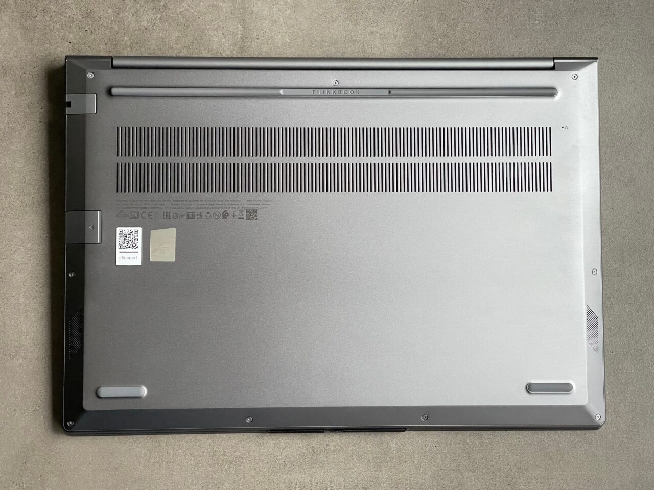 Spód laptopa umieszczonego na szarym tle, z widocznymi otworami wentylacyjnymi, śrubami, naklejkami z informacjami i numerem seryjnym oraz gumowymi nóżkami.