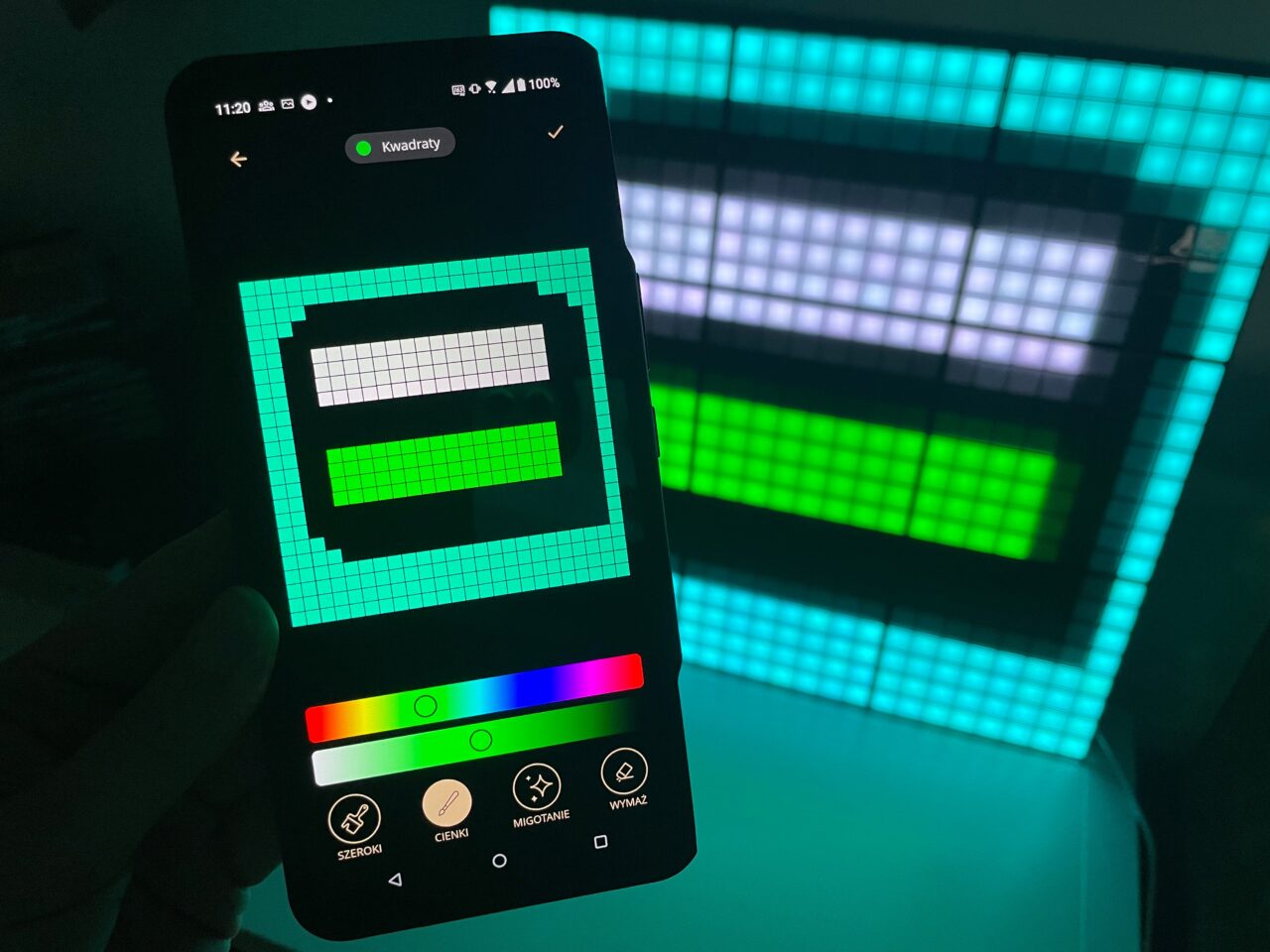 Dłoń trzymająca smartfon z otwartą aplikacją do tworzenia wzorów pikselowych nałączonym ekranem, pokazującym prostokątny wzór, odzwierciedlony na większym, podświetlanym panelu LED w tle.