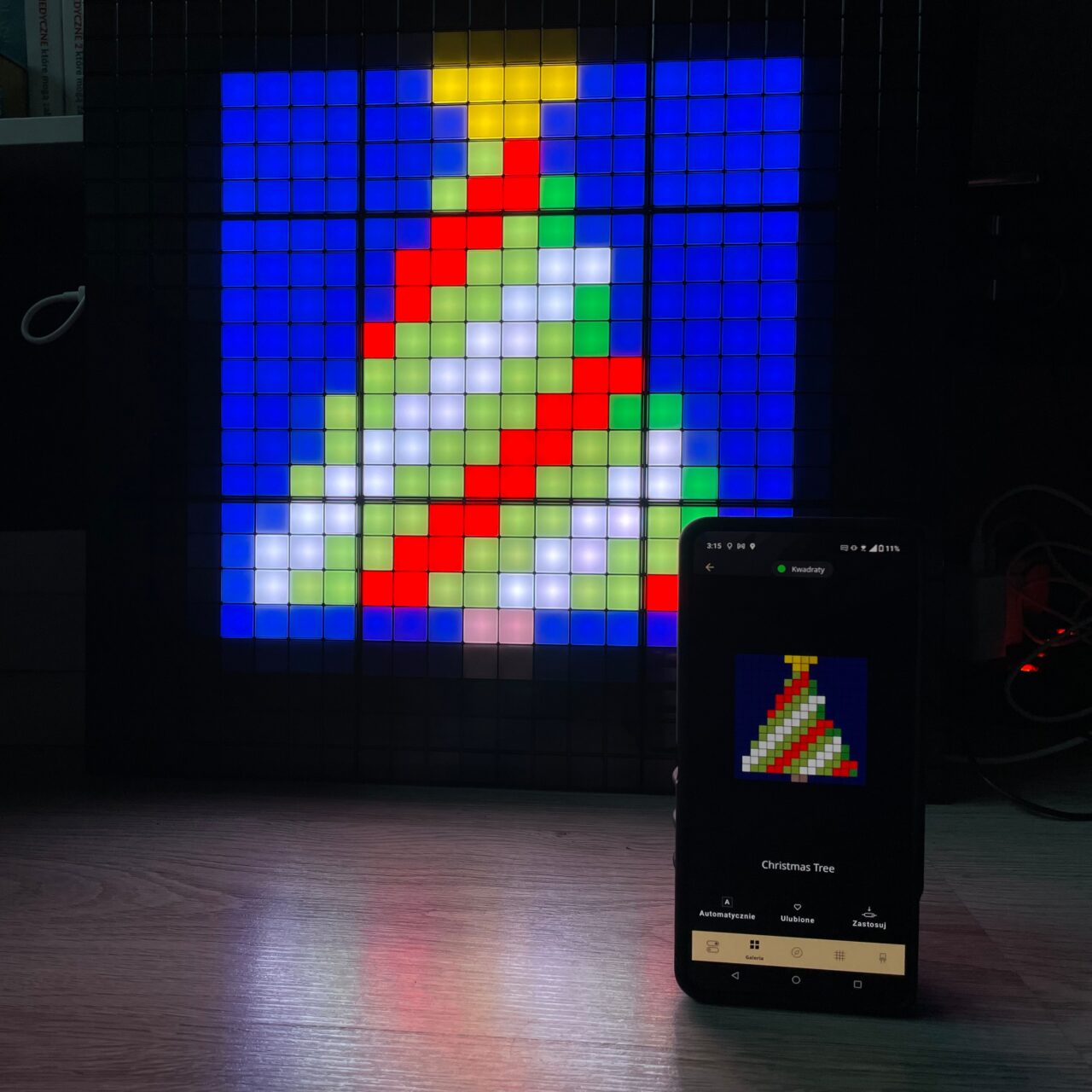 Inteligentna tablica świetlna na ścianie wyświetlająca pixel art choinki z towarzyszącym obrazem tej samej choinki na ekranie smartfona, umieszczonego pionowo na podłodze.