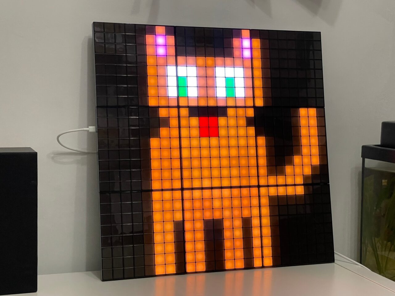 Cyfrowa, pikselowa reprezentacja kota w pomarańczowej i czarnej kolorystyce na kwadratowym, podświetlanym panelu LED zamontowanym na ścianie.