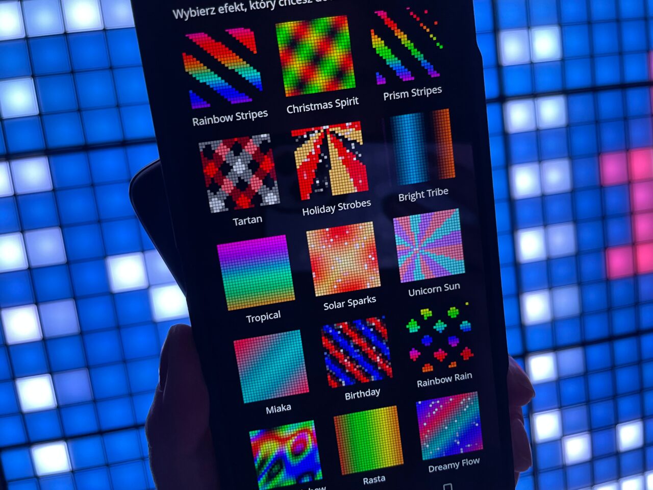 Ekran smartfona wyświetlający różne wzory świetlne z opisami w aplikacji, trzymany na tle świecącej ściany z kwadratowymi panelami LED.
