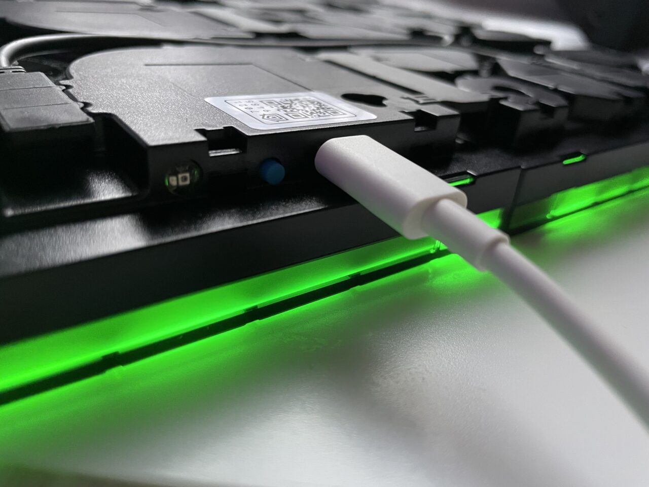 Częściowo otwarta czarna klawiatura laptopa z podświetleniem LED w odcieniu zielonym oraz białym kablem ładowania podłączonym do boku.