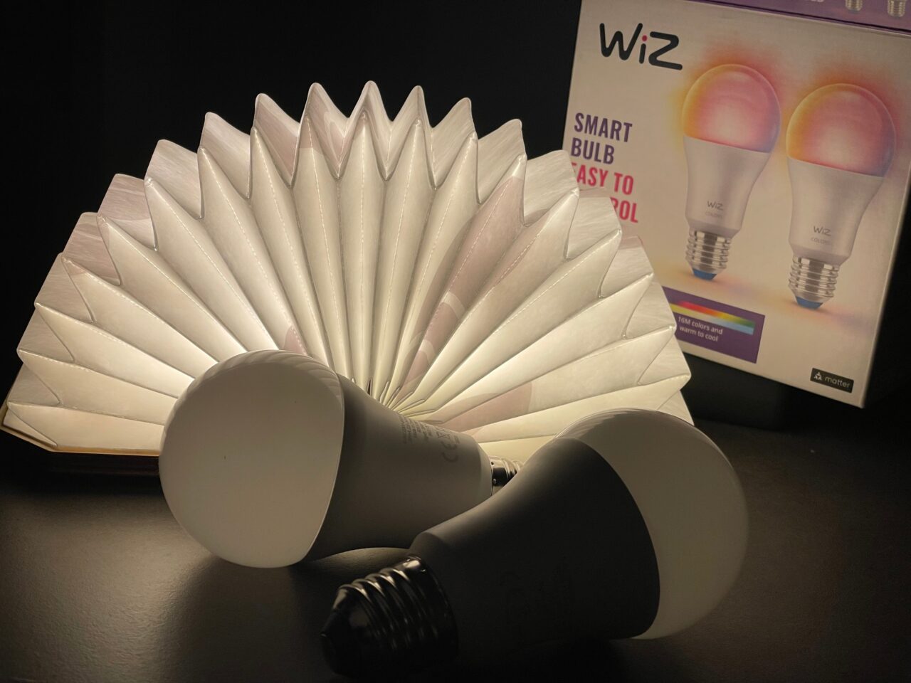 Dwie inteligentne żarówki LED firmy WiZ leżące na ciemnej powierzchni obok rozłożonej lampy książkowej i opakowania produktu.