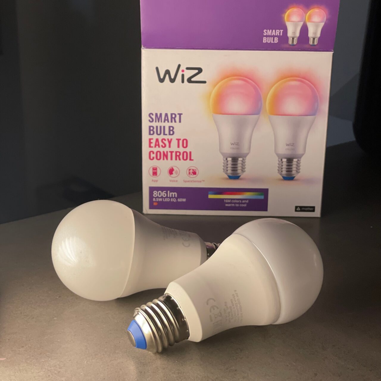 Dwie inteligentne żarówki LED marki WiZ leżące na blacie z opakowaniem w tle, które pokazuje te same żarówki oraz informacje o łatwości sterowania i specyfikacji produktu.