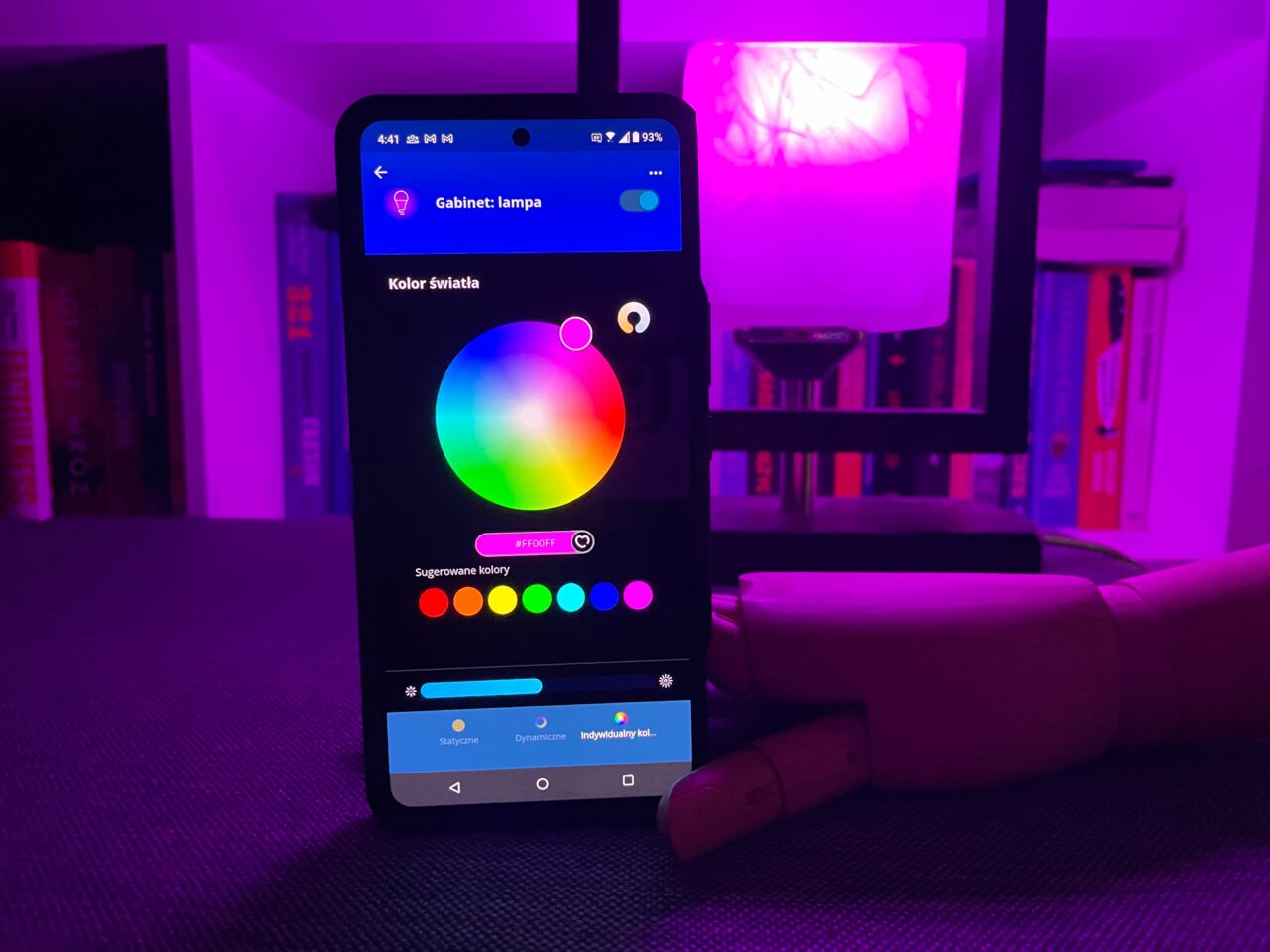 Smartfon wyświetlający aplikację do sterowania kolorowym oświetleniem w pomieszczeniu z fioletowym światłem w tle.