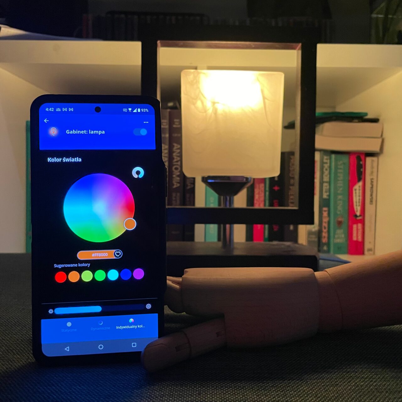 Inteligentny telefon wyświetlający aplikację do sterowania kolorami lampy, umieszczony przy włączonej lampce na tle półki z książkami.