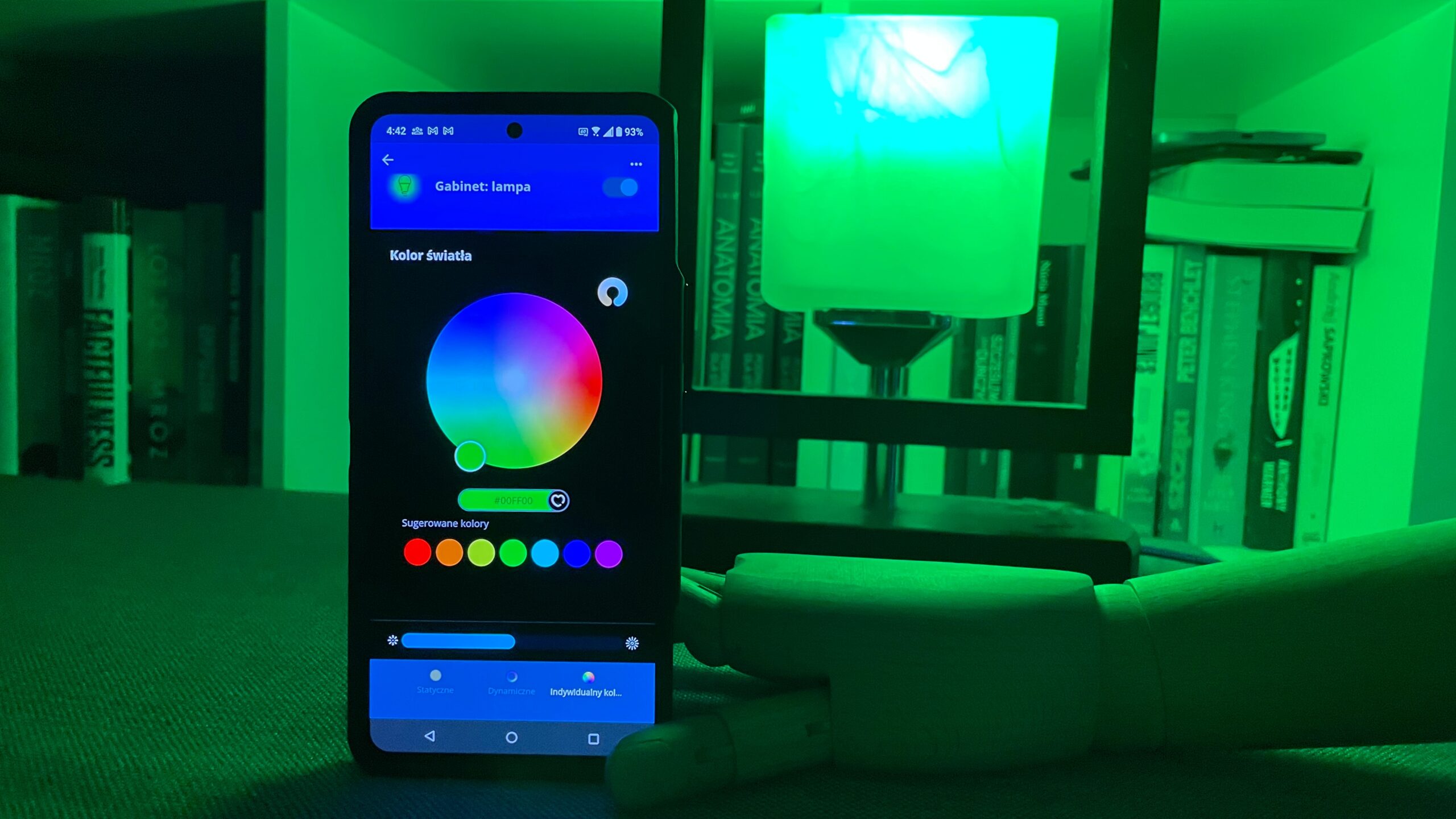Smartfon wyświetlający aplikację do sterowania kolorowym oświetleniem w zielonym oświetleniu pokoju z półkami z książkami w tle.