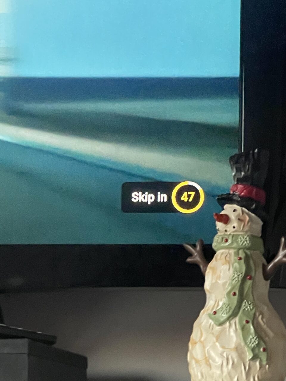 Figurka bałwana stoi przed ekranem telewizora z wyświetlaczem "Skip in 47" w prawym górnym rogu w usłudze Google, YouTube.