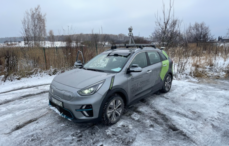 Samochód z napisem "SELF-DRIVING VEHICLE" zaparkowany na oblodzonej nawierzchni z zimowym krajobrazem w tle.