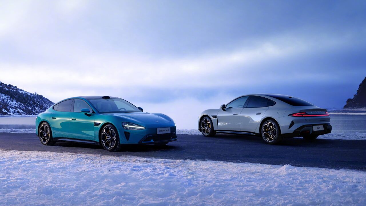 dois carros elétricos Xiaomi SU7, um cinza e outro azul, passando um pelo outro na estrada em uma paisagem de inverno