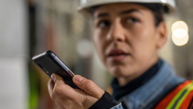 Pracownik w kasku ochronnym patrzy na telefon komórkowy, w tle nieostry obraz wnętrza przemysłowego.