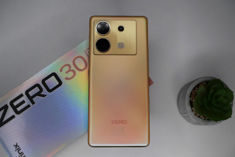 Złoty smartfon z potrójnym aparatem umieszczony na biurku obok pudełka z napisem "ZERO 30" i doniczką z zieloną sukulentem.