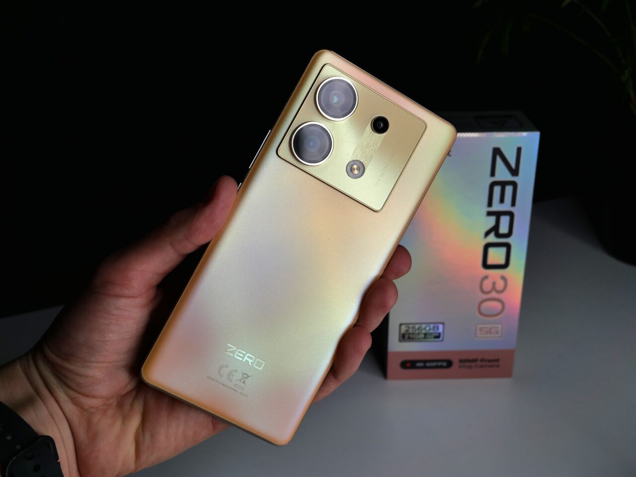 Ręka trzymająca złoty smartfon z trzema obiektywami aparatu i logo na plecach, obok opakowanie z napisem "ZERO 30 5G".