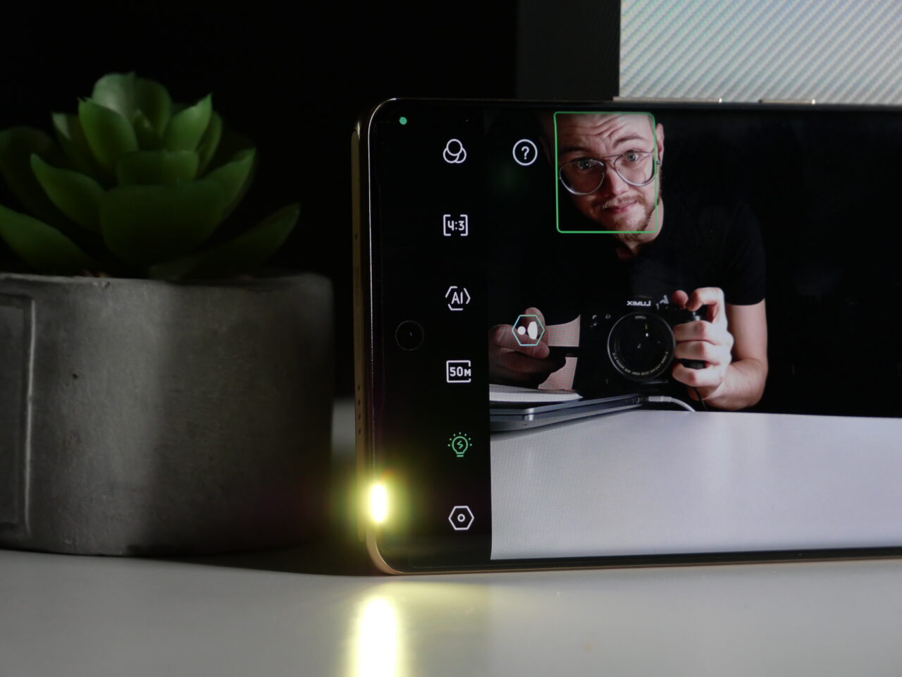 Człowiek robiący selfie za pomocą smartfona, który leży na biurku obok doniczki z zieloną rośliną; twarz człowieka jest wykadrowana na ekranie smartfona.
