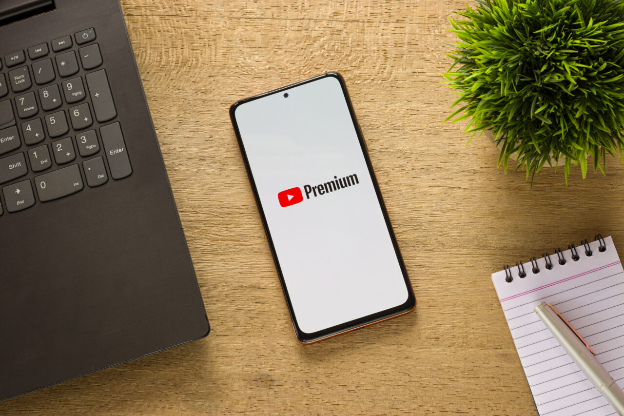 Smartfon z logo YouTube Premium, usługą kasującą reklamy na YouTube, na ekranie. Urządzenie leży na biurku obok laptopa, sztucznej rośliny i notatnika z długopisem.
