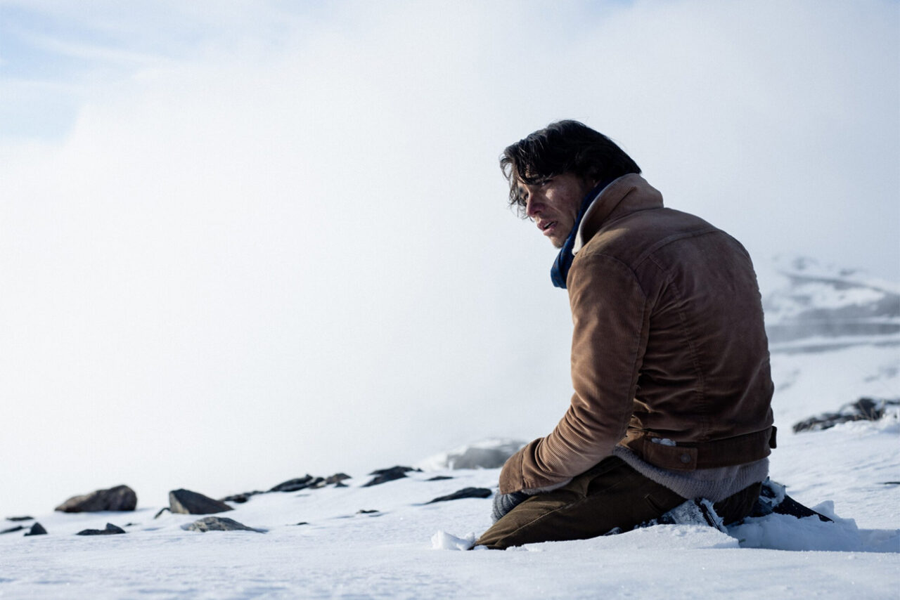 Mężczyzna w brązowej kurtce siedzi na śniegu, wyglądając na przemarzniętego i wyczerpanego, w tle zamglone górskie krajobrazy. Kadr z filmu Śnieżne bractwo od Netflix