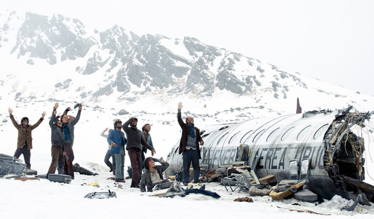 Grupa ocalałych z katastrofy lotniczej, machających rękami i wołających o pomoc, wokół wraku samolotu na śnieżnym stoku górskim. Kadr z filmu Śnieżne bractwo od Netflix