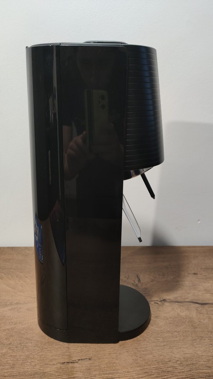 Czarny, cylindryczny SodaStream Terra stojący na drewnianym blacie, odbijający w swojej błyszczącej powierzchni odbicie osoby robiącej zdjęcie smartfonem.