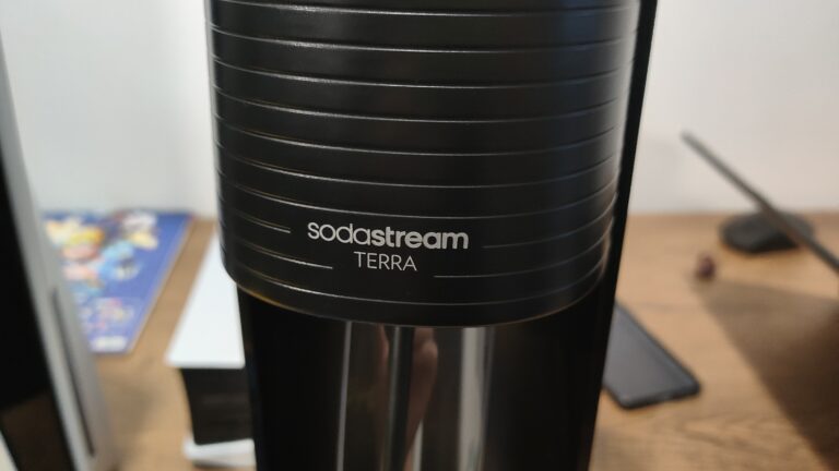 Część czarnego urządzenia SodaStream TERRA z widocznym logo marki, na tle rozmytego wnętrza z deską, telefonem komórkowym i innymi przedmiotami.