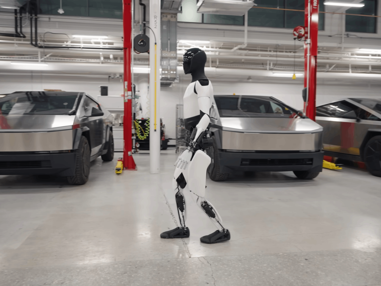Robot humanoidalny Tesla Optimus w czarno-białej kolorystyce chodzi w garażu z zaparkowanymi pojazdami Tesla Cybertruck