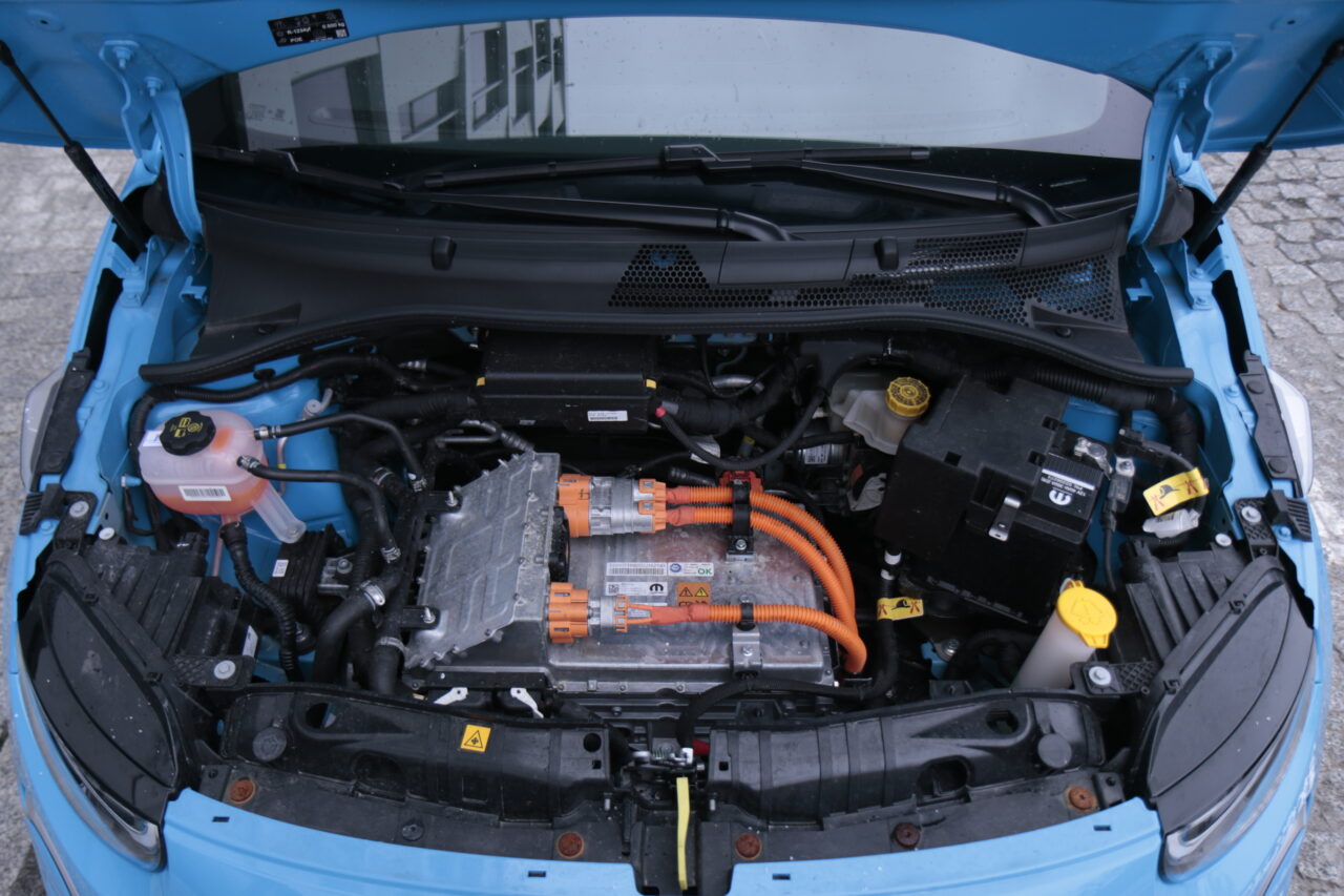 Podniesiona maska niebieskiego samochodu elektrycznego ujawniająca baterię i inne składniki układu napędowego.