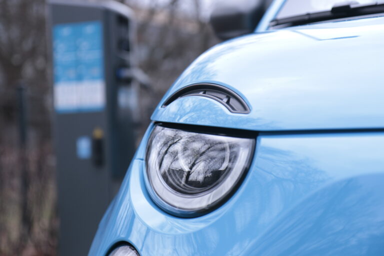 Niebieski samochód elektryczny - elektrykiem na święta - z widoczną refleksją gołych drzew w okrągłym reflektorze, w tle nieostrze widoczna ładowarka do pojazdów elektrycznych.