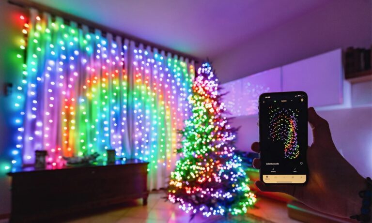 Pokój z choinką ozdobioną w świąteczne lampki i ścianą z kolorowymi, pionowymi światełkami, sterowanymi za pomocą aplikacji na smartfonie w ręce osoby.