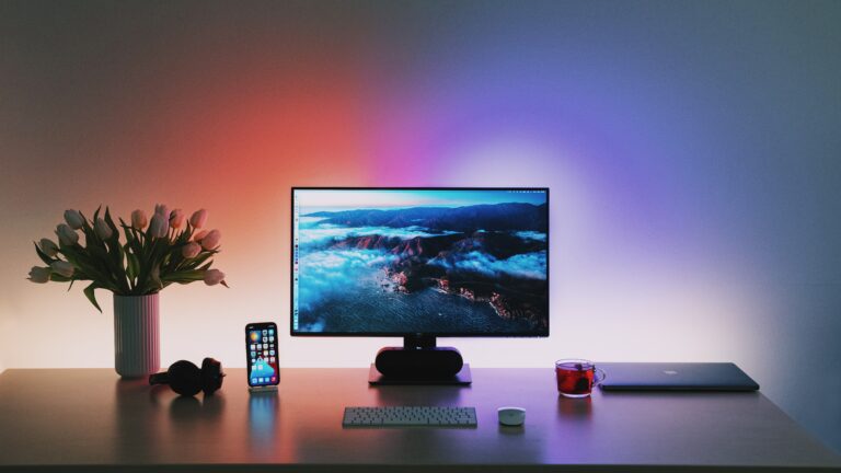 Zestaw biurkowy z monitorem na środku wyświetlającym krajobraz, obok słuchawki, smartfon, laptop, kubek z czerwonym napojem i wazon z tulipanami, podświetlenie LED zmieniające kolor na ścianie.