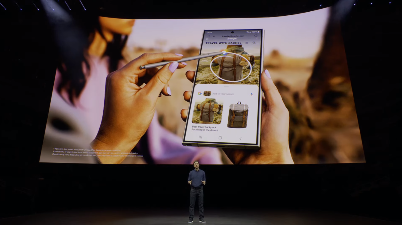 Prezentacja na scenie z dużym ekranem pokazującym dłonie trzymające smartfon i korzystające z funkcji wyszukiwania obrazami, w tle widz mały ekran i stojącą osobę.