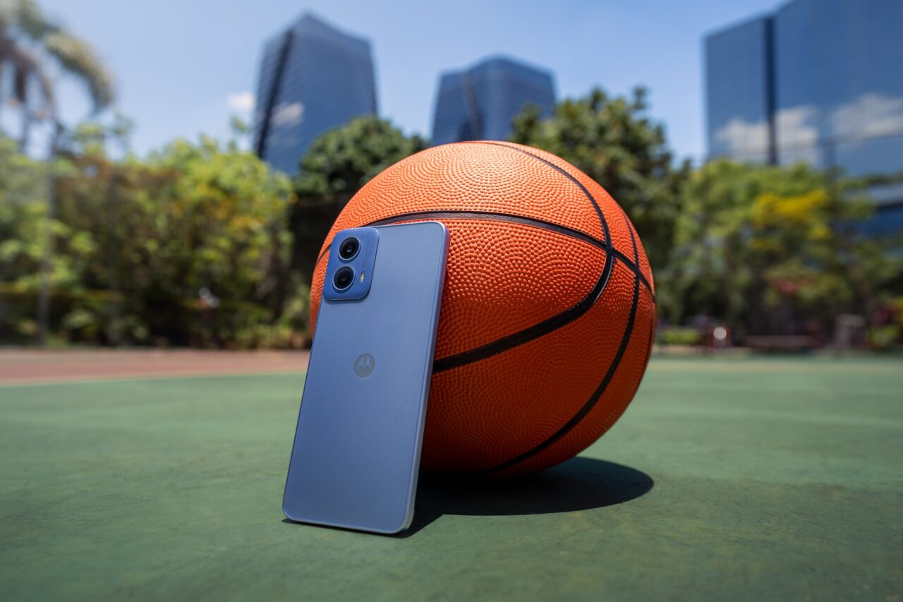 Piłka do koszykówki i smartfon marki Motorola oparte o siebie na zewnątrz na tle kortu do koszykówki i wysokich, nowoczesnych budynków.