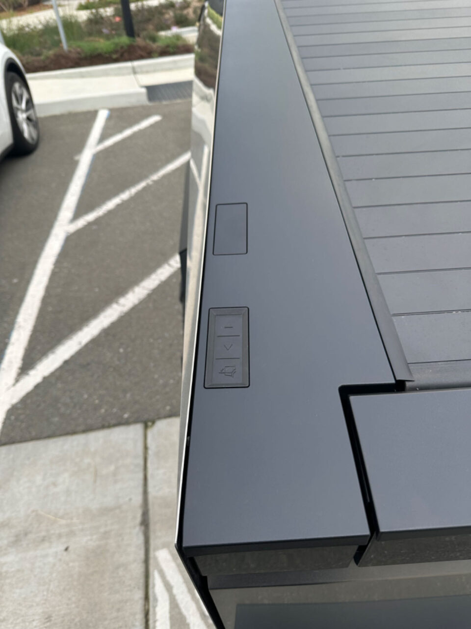 Widok na panel sterowania windą zewnętrzną z przyciskami do wyboru piętra, przy nieostrych liniiach oznakowania parkingu w tle.