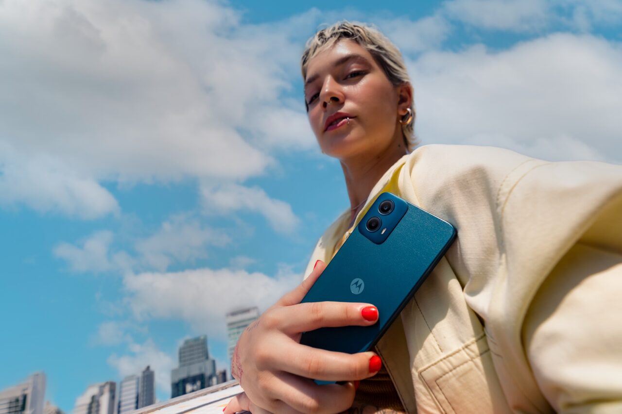 Kobieta trzymająca smartfon z tyłu, z widokiem na niebieskie niebo i zabudowania miasta w tle.