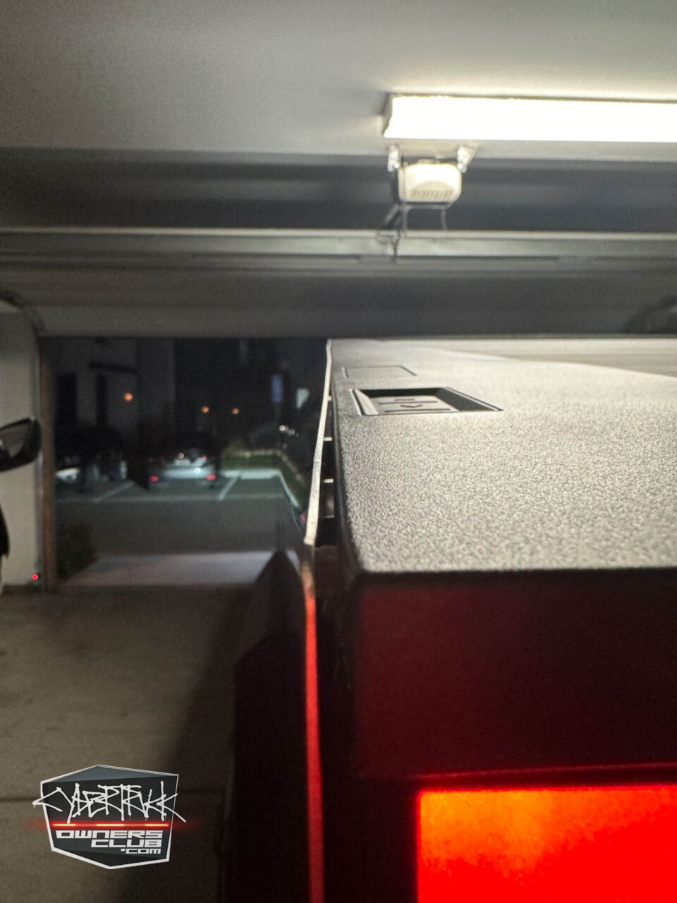 Zdjęcie z perspektywy wnętrza garażu, pokazujące fragment czerwonego tylnego światła samochodu i górną część otwartej garażowej bramy, przez którą widać parkujące na zewnątrz pojazdy; w tle świeci jasna lampa garażowa.