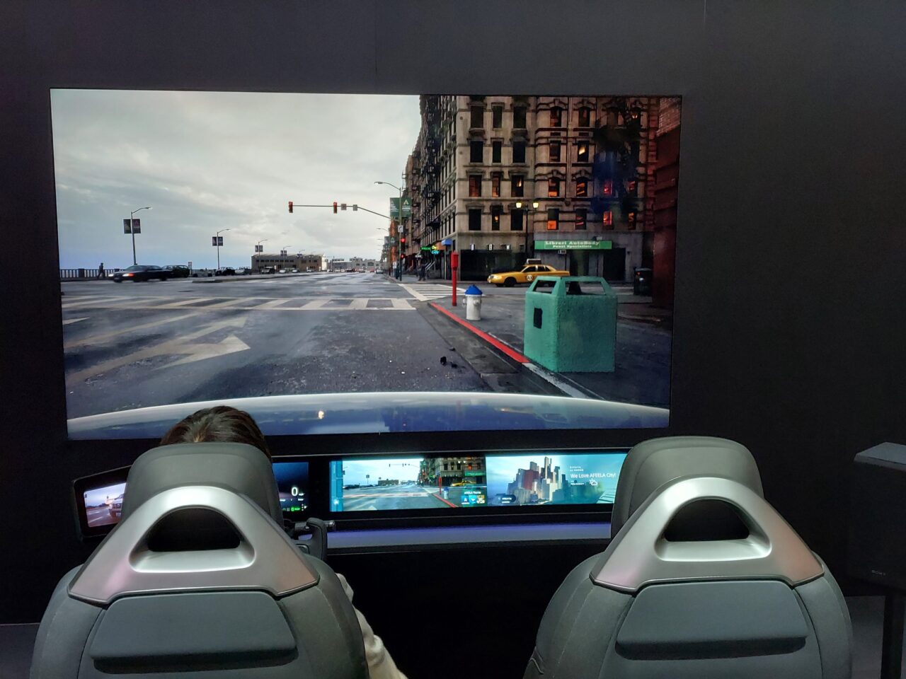 Tylna część głowy osoby siedzącej w symulatorze jazdy z panoramycznym wyświetlaczem przedstawiającym miejską scenę z ulicą i światłami drogowymi.