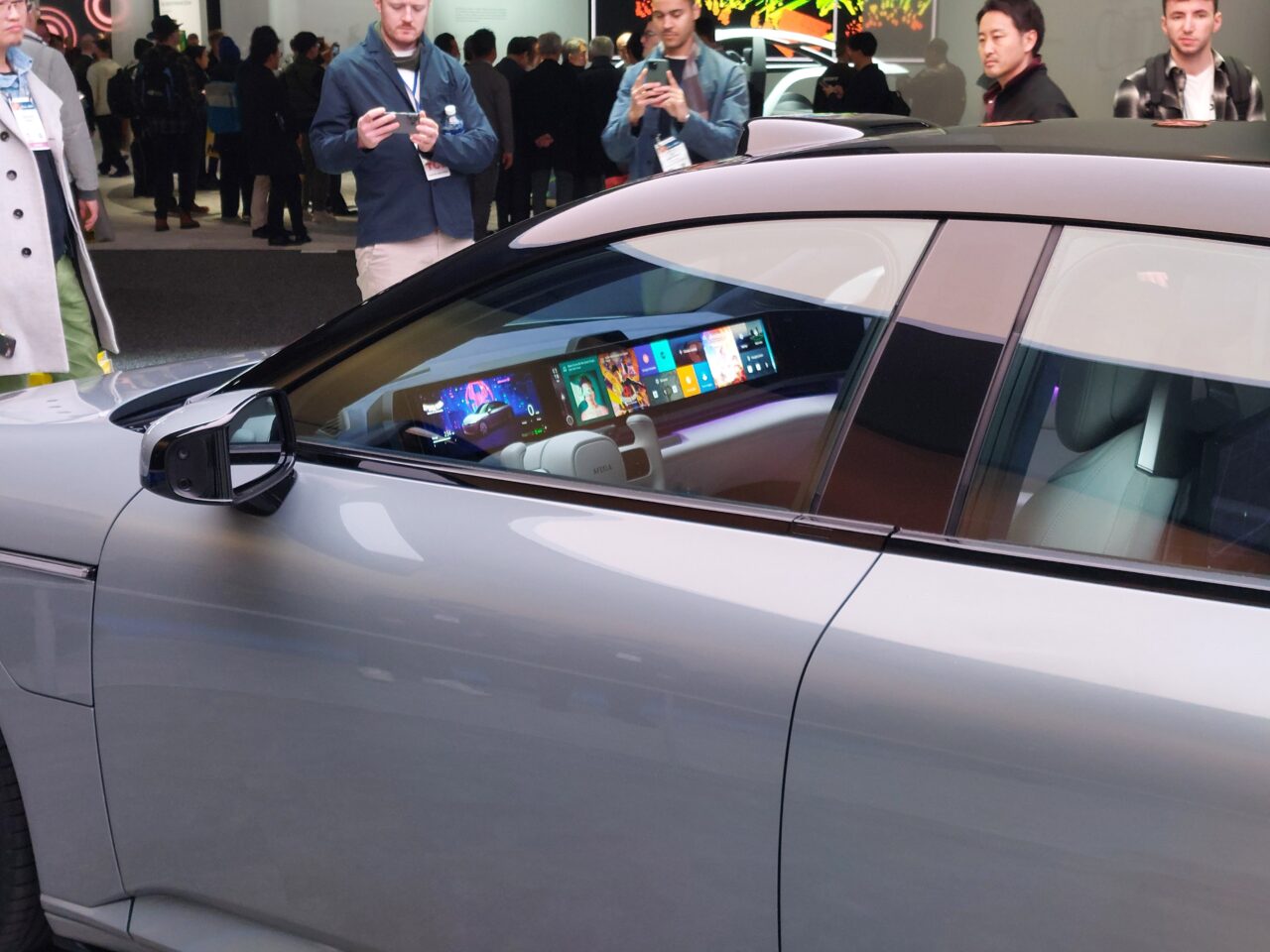 Samochód z zaawansowanym systemem multimedialnym widoczny przez boczną szybę, na tle ludzi na wystawie technologicznej.