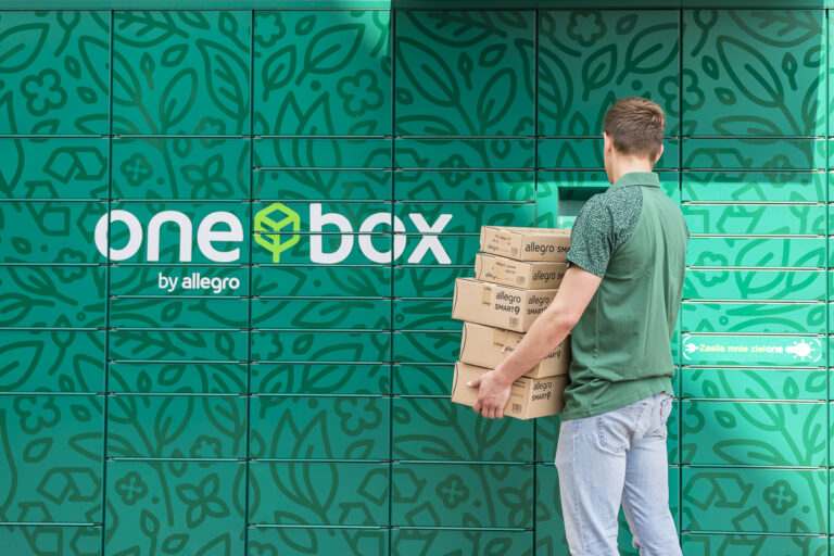 Mężczyzna trzymający stos kartonowych pudełek przed zieloną ścianą paczkomatu "one box by allegro".