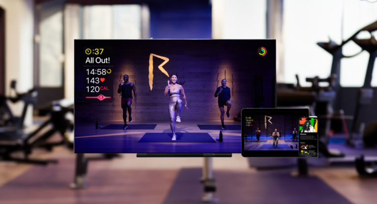 Trzy osoby uczestniczące w zajęciach fitness transmitowanych na żywo na duży ekran w siłowni oraz na tablet na pierwszym planie.