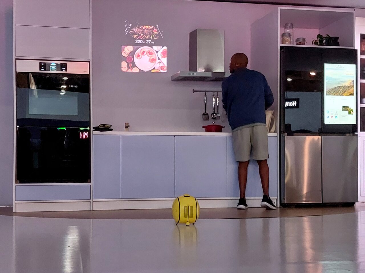 Mężczyzna stoi przy blacie kuchennym; nowoczesne szafki i lodówka z ekranem; na podłodze żółty robot Samsung Ballie.