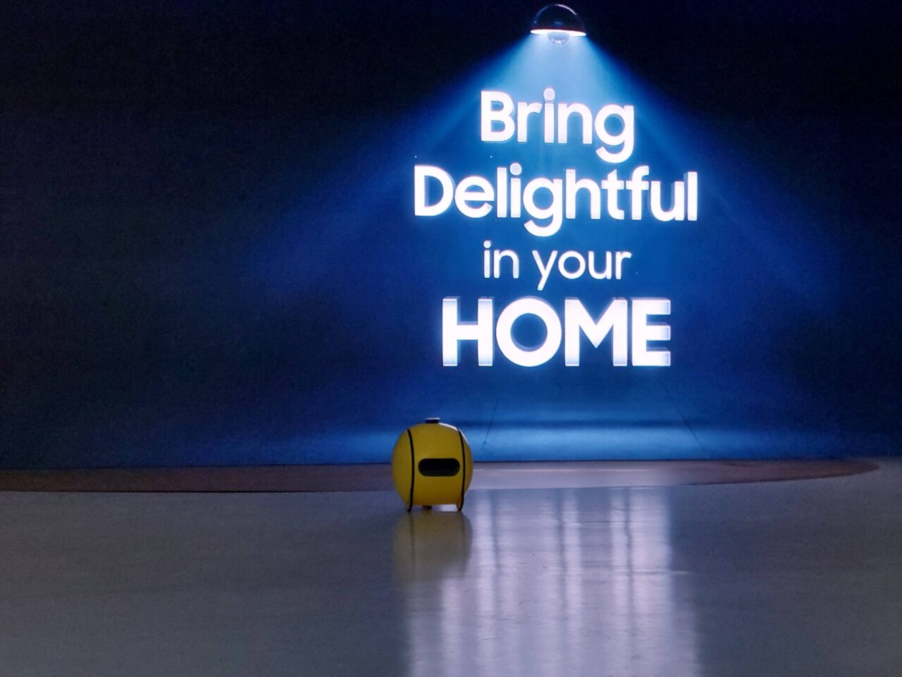 Przedstawienie małego, żółtego robota Samsung Ballie stojącego na podłodze w pomieszczeniu z niebieskim oświetleniem i napisem na ścianie "Bring Delightful in your HOME" w tle.
