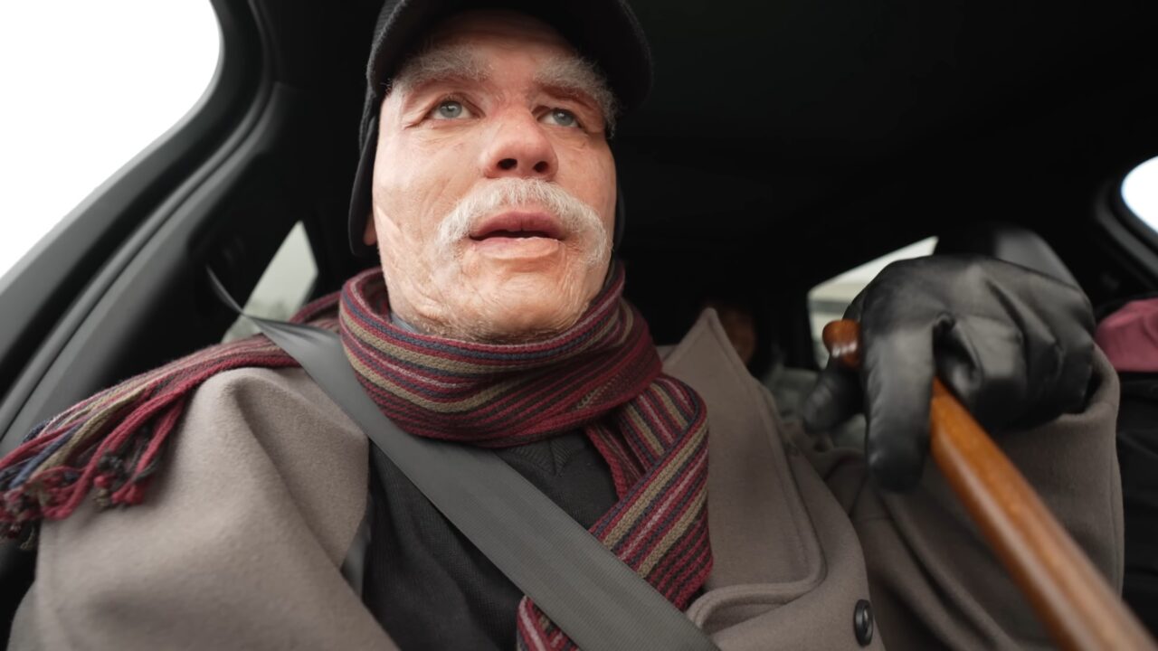 Budda na najnowszym filmie. Starszy mężczyzna z białym wąsem, ubrany w beret, szalik, płaszcz i rękawiczki, siedzący na przednim fotelu samochodu, patrzący w górę z zamyślonym wyrazem twarzy.