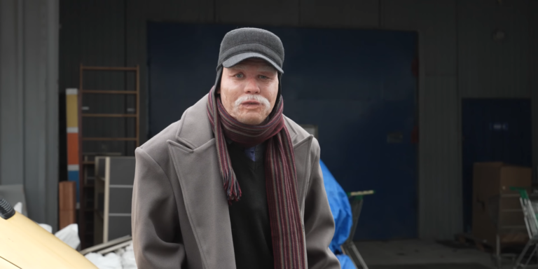 Mężczyzna w średnim wieku z kapturem i szalikiem stojący na tle przemysłowego budynku.