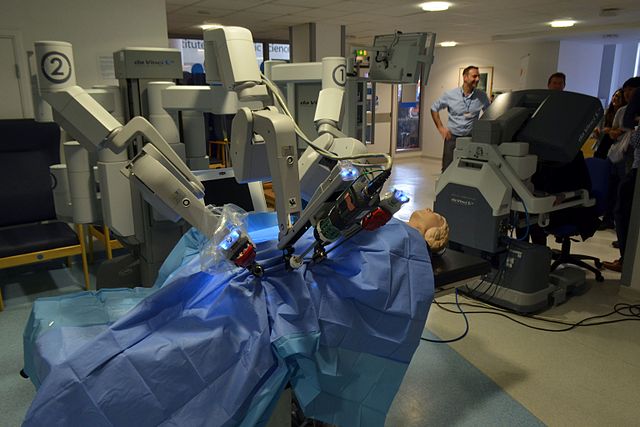 Robotyzacja medycyny jako przyszłość opieki zdrowotnej. Robot chirurgiczny typu da Vinci podczas pokazu, z manekinem pacjenta na stole operacyjnym, w tle personel medyczny i obserwatorzy.
