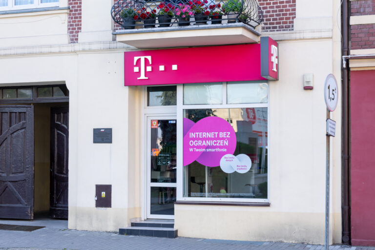 Widok na front salonu firmy telekomunikacyjnej T-Mobile z logo na znaku i okleinie promującej "Internet bez ograniczeń" na witrynie sklepu.