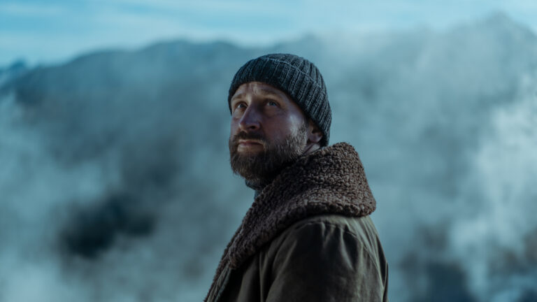 Mężczyzna w czapce i kurtce patrzy w dal, w tle rozmyte górskie szczyty i niebo. Kadr z serialu Forst dla Netflix.