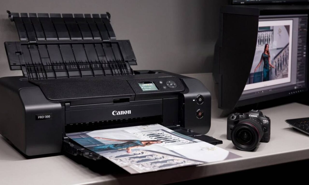 Profesjonalna drukarka Canon PRO-300 drukująca kolorowe zdjęcie na biurku przy komputerze, obok aparatu fotograficznego Canon EOS.