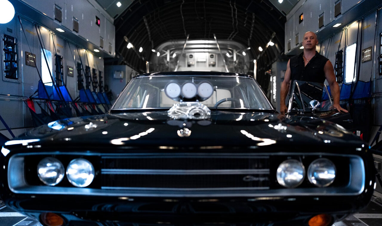 Czarny klasyczny samochód Dodge Charger z wystającym silnikiem w ładowni samolotu, za nim stoi mężczyzna oparty o maszynę. Zdjęcie z filmyu Fast X ilustrujące artykuł o kontynuacji pt. Szybcy i wściekli 11